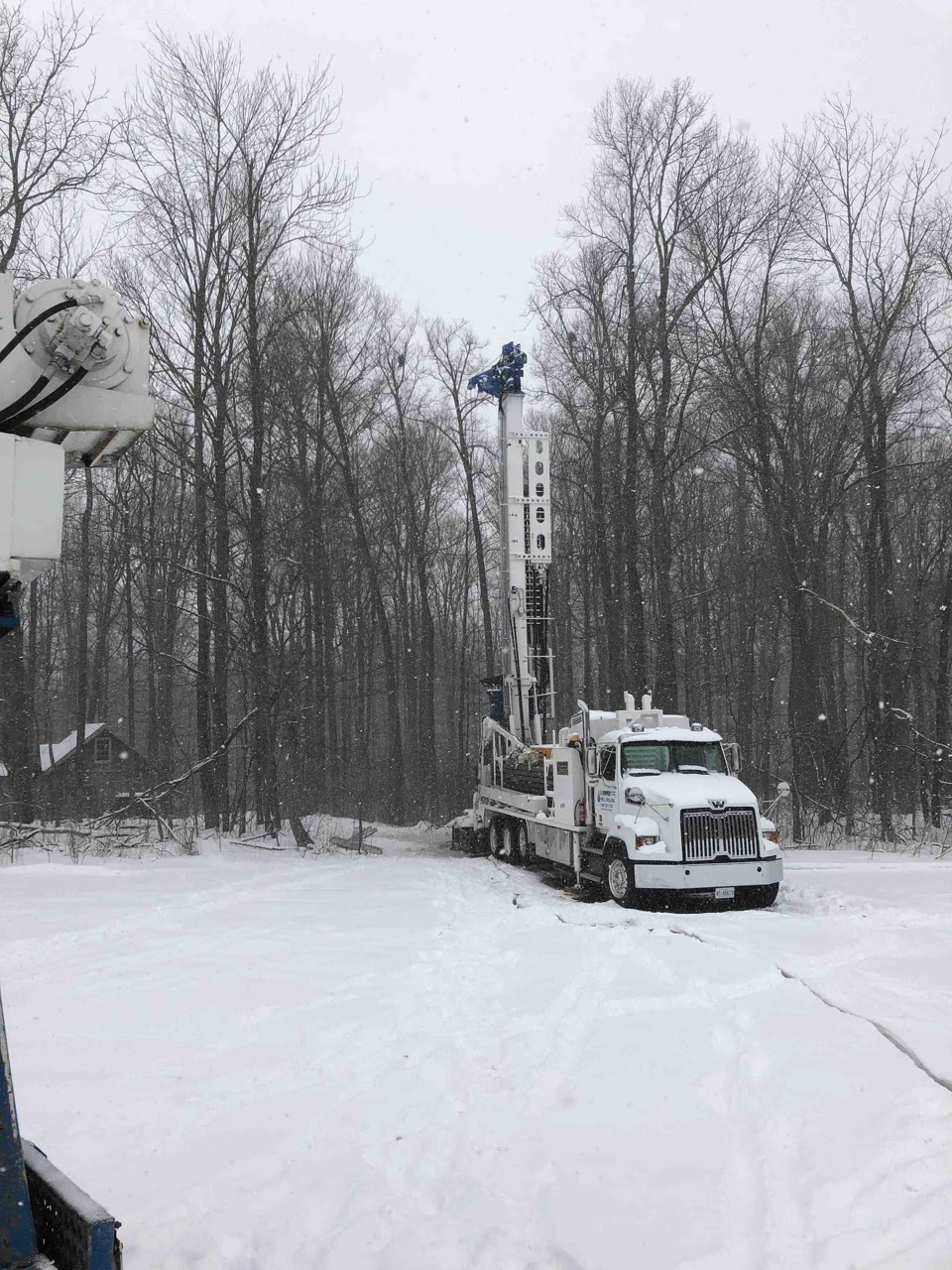A Hopper drill rig with mast raised drilling in falling snow at a treeline