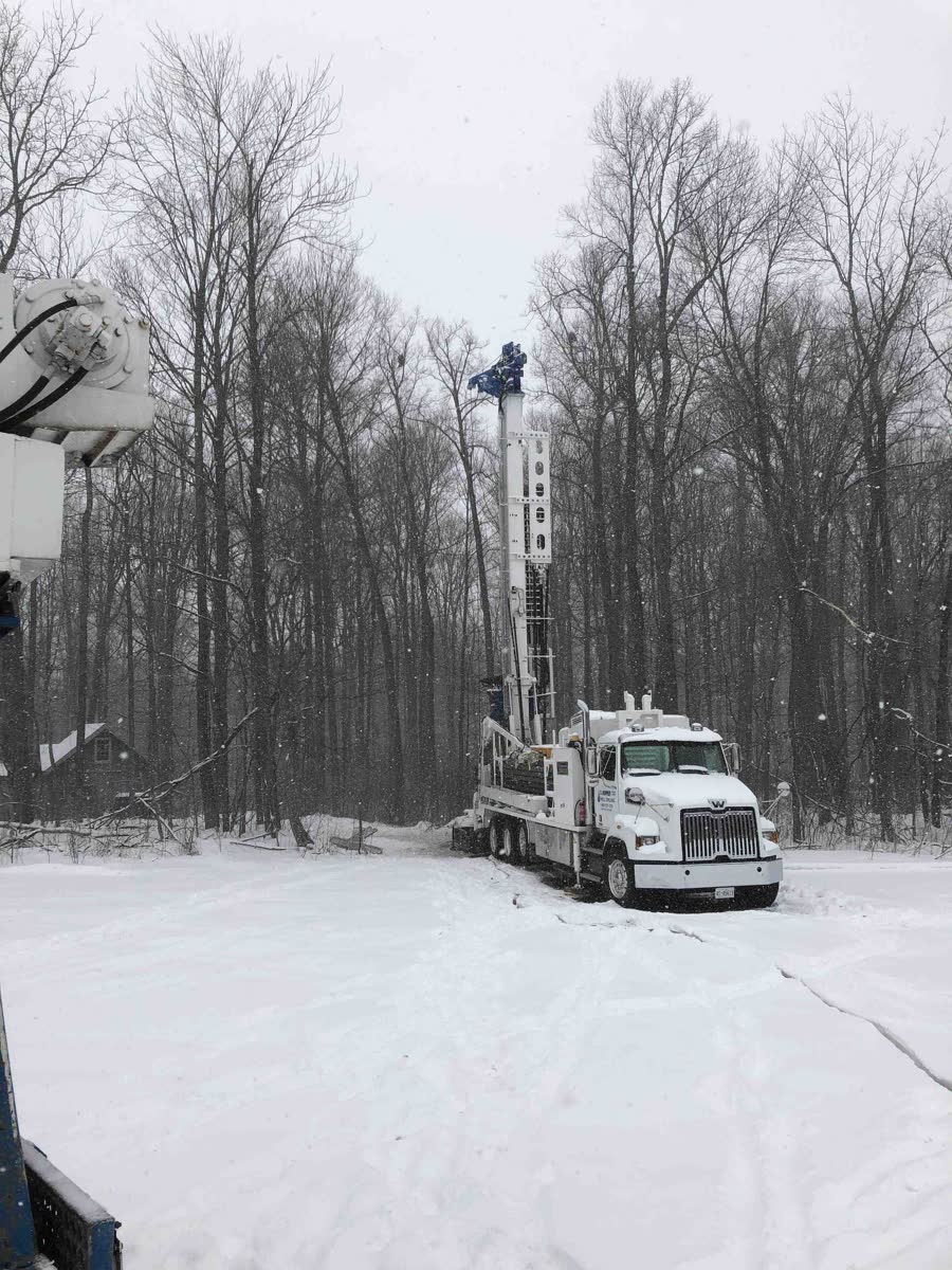 A W.D. Hopper drill rig set up at the edge of a snowy, wooded property, mast raised